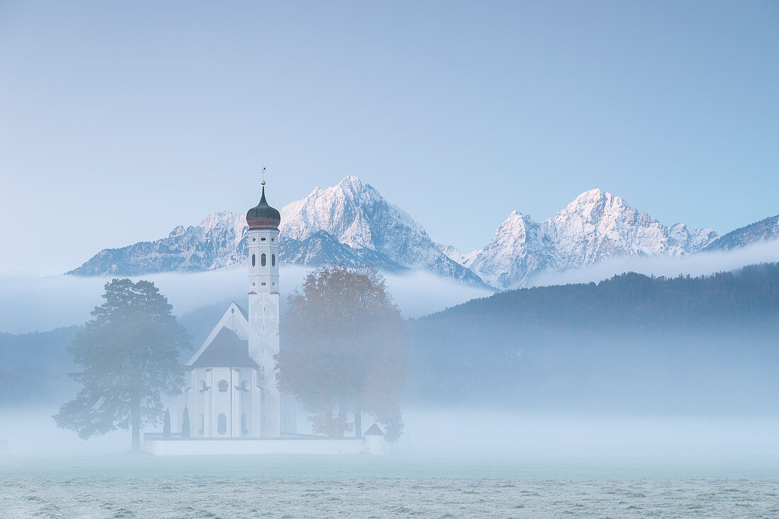 The autumn fog at sunrise frames St. Coloman Church surrounded by snowy peaks, Schwangau, Bavaria, Germany, Europe