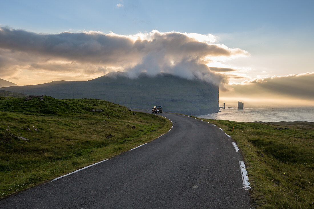 Auto folgt einer kurvenreichen Straße bei Sonnenuntergang, Klippen und Ozean im Hintergrund, Eidi, Eysturoy, Faroe Islands, Dänemark