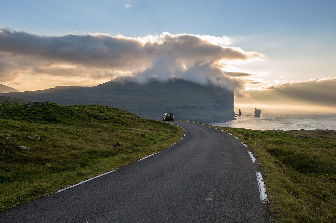 CAR FOLLOWING A WINDING ROAD AT SUNSET, CLIFFS AND OCEAN IN THE BACKGROUND, EIDI, EYSTUROY, FAROE ISLANDS, DENMARK