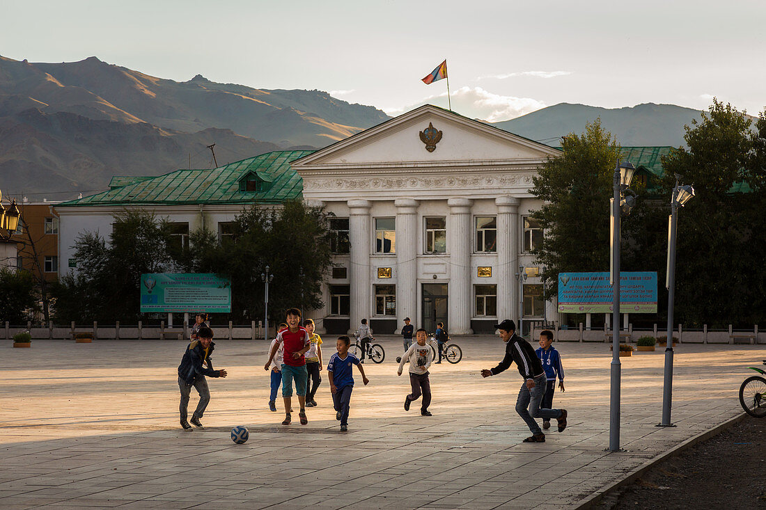 Kinder spielen Fussball auf dem Hauptquadrat vor einem Amtsgebäude in der Stadt Ulgii und den umgebenden Bergen, Provinz Bayan-Olgii, Mongolei