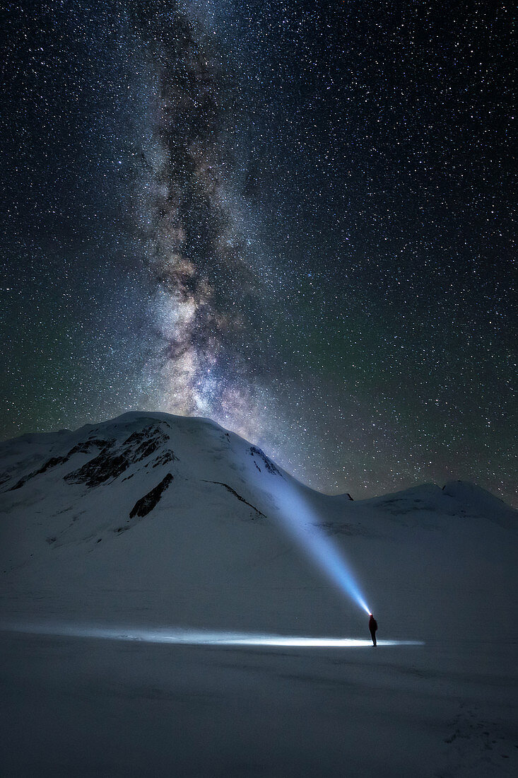 Bergsteiger betrachtet den Nachthimmel und die Milchstrasse, während er den Berg Khuiten mit seiner Scheinlampe beleuchtet, Altai, Tavan Bogd Massif, Provinz Bayan-Olgii, Mongolei