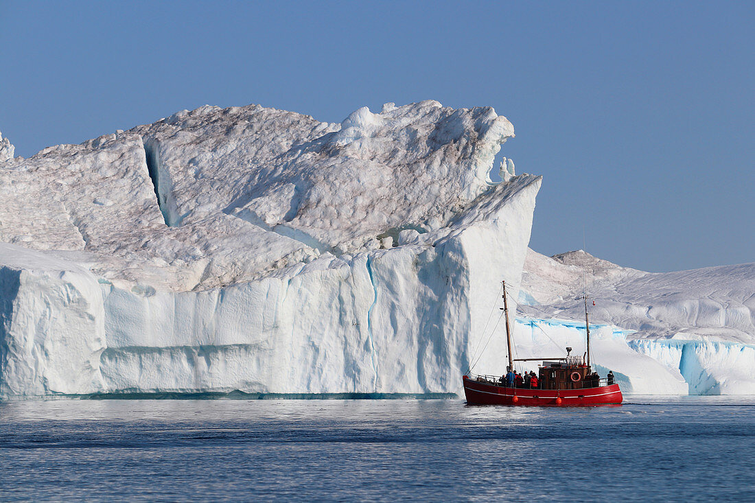 Disco Bay in West Greenland; Icebergs in the Kangia Icefjord near Ilulissat; red tour boat with visitors approaches an iceberg; Steep walls and demolition areas; Sun and cloudless sky;