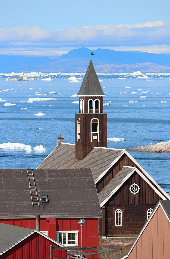 Zionskirche in Ilulissat; Disco Bay in West Greenland; Church covered with brown painted wood; white window frames and roof edges; in the foreground sections of wooden houses; in bright red and light brown; behind the church is the Disko Bay with icebergs and ice floes; mountain ranges in the background