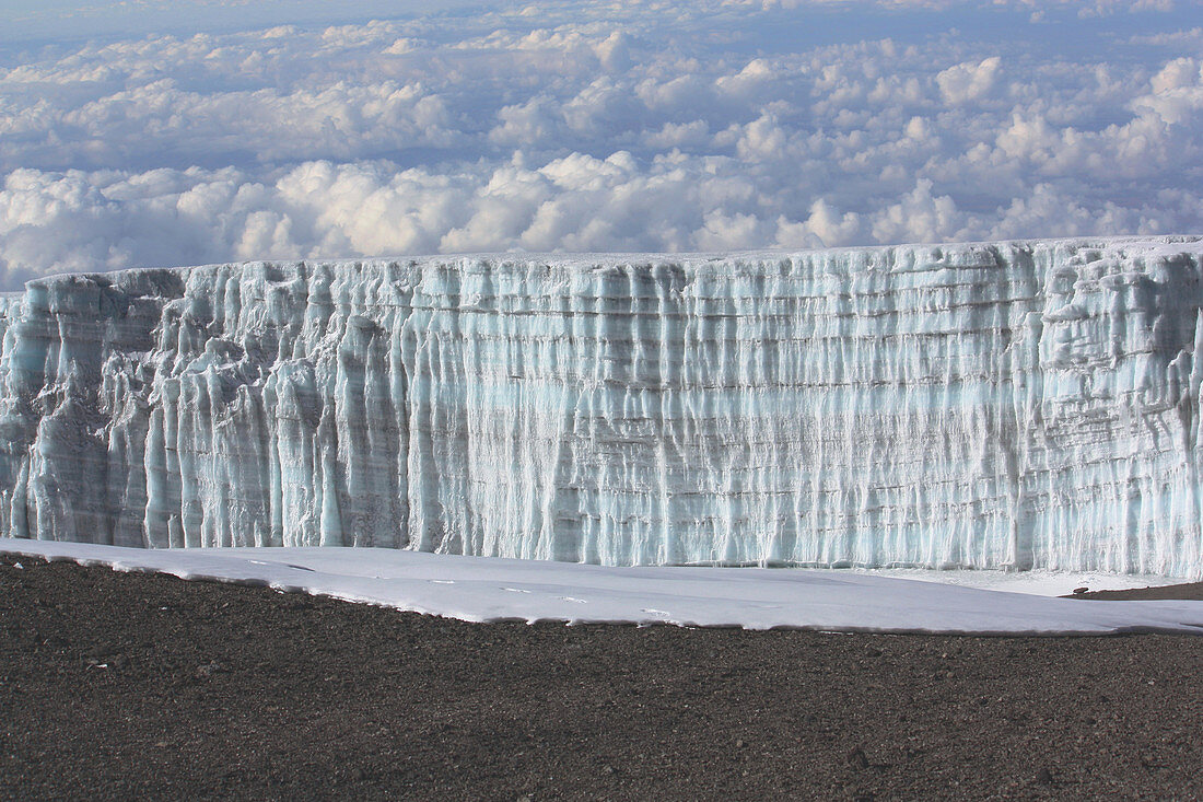 Blick vom Gipfel des Kilimandscharo auf die Gletscherwand, Lavageröll, teilweise mit Schnee bedeckt, dahinter Wolkenmeer