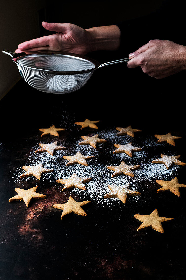 High angle close up of person dusting freshly baked star-shaped cookies with icing sugar.