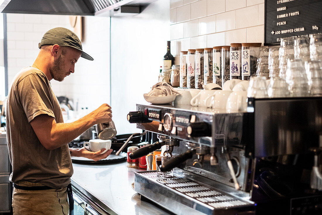 Bärtiger Mann mit Baseballmütze bereitet Cappuccino an der Espressomaschine zu