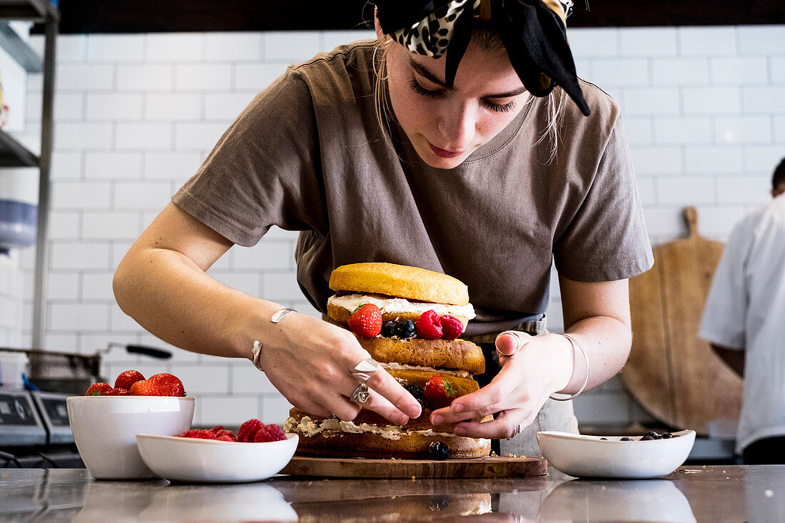A cook working in a commercial kitchen assembling a layered sponge cake with fresh fruit.