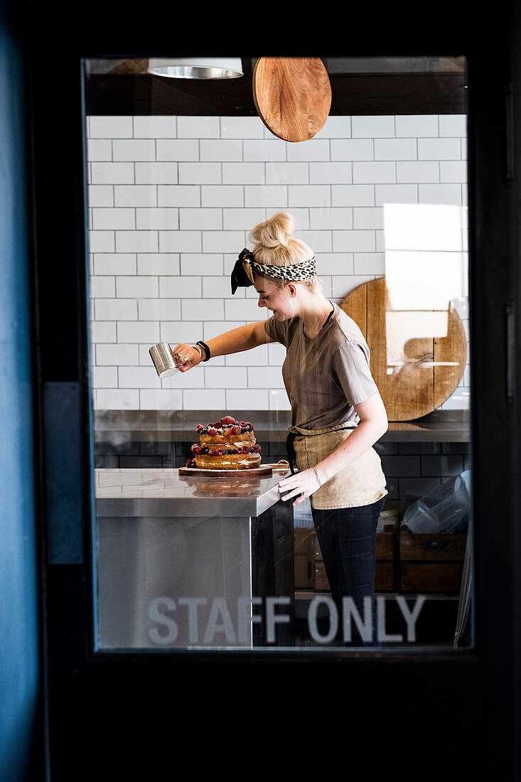 Blick durch eine mit 'nur für Personal' gekennzeichnete Tür auf Köchin, die einen Schichtkuchen mit frischem Obst mit Puderzucker bestäubt
