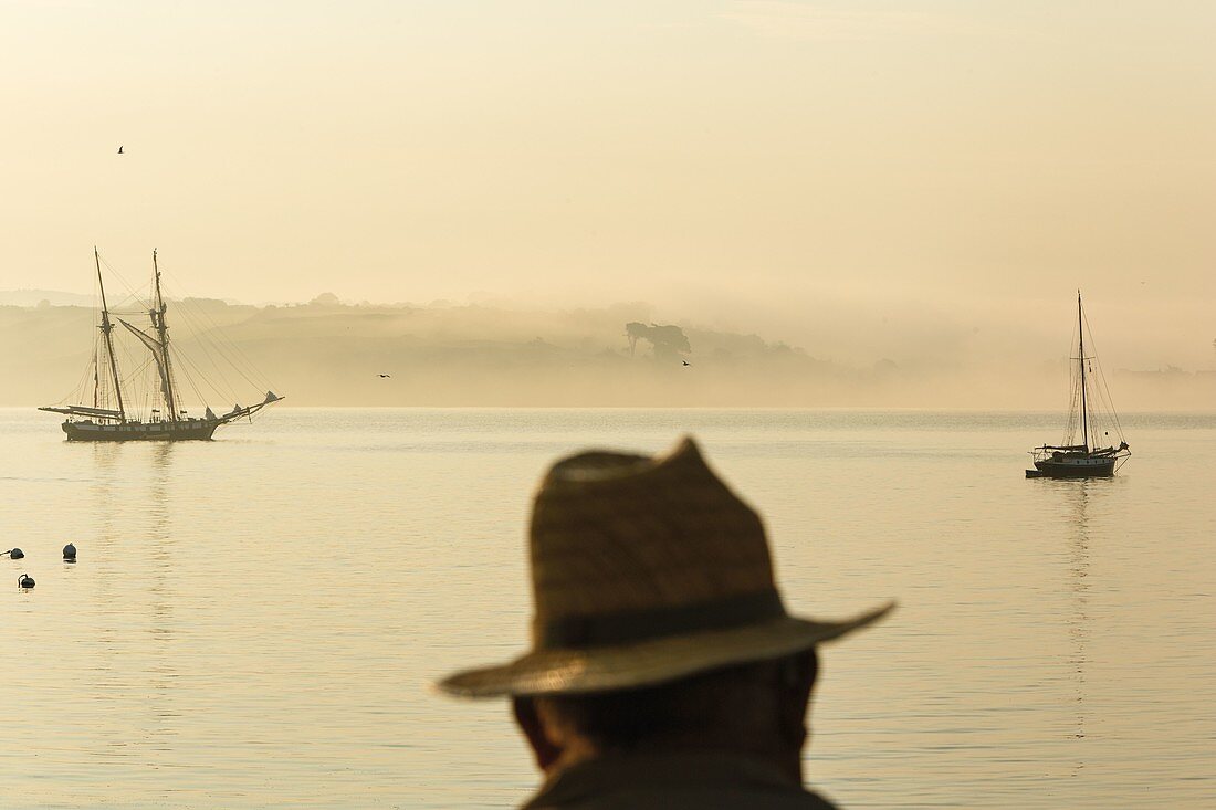 France, Finistere, Douarnenez, scene of life, man at dawn facing a two-masted and small coastal fishing boat with landscape in the mist