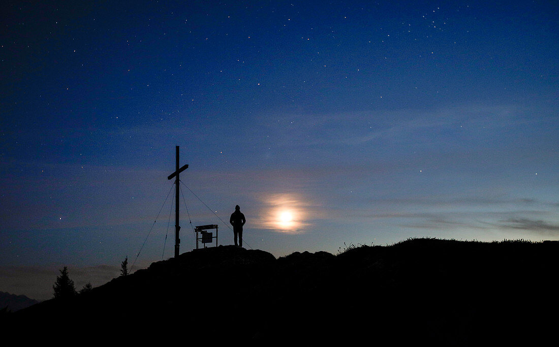 Person at the summit cross of the Jochberg at night with clouds, stars and moon