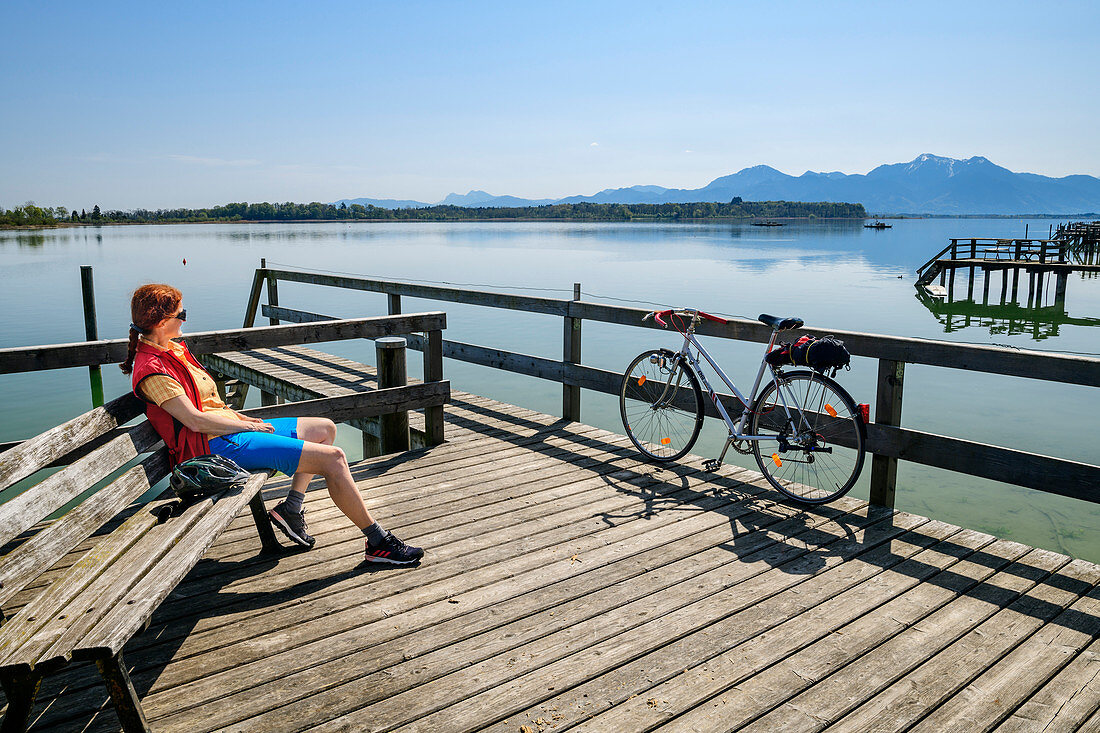 Frau beim Radfahren sitzt an Steg und blickt auf Chiemsee, Hochgern im Hintergrund, Chiemseeradweg, Chiemgau, Oberbayern, Bayern, Deutschland