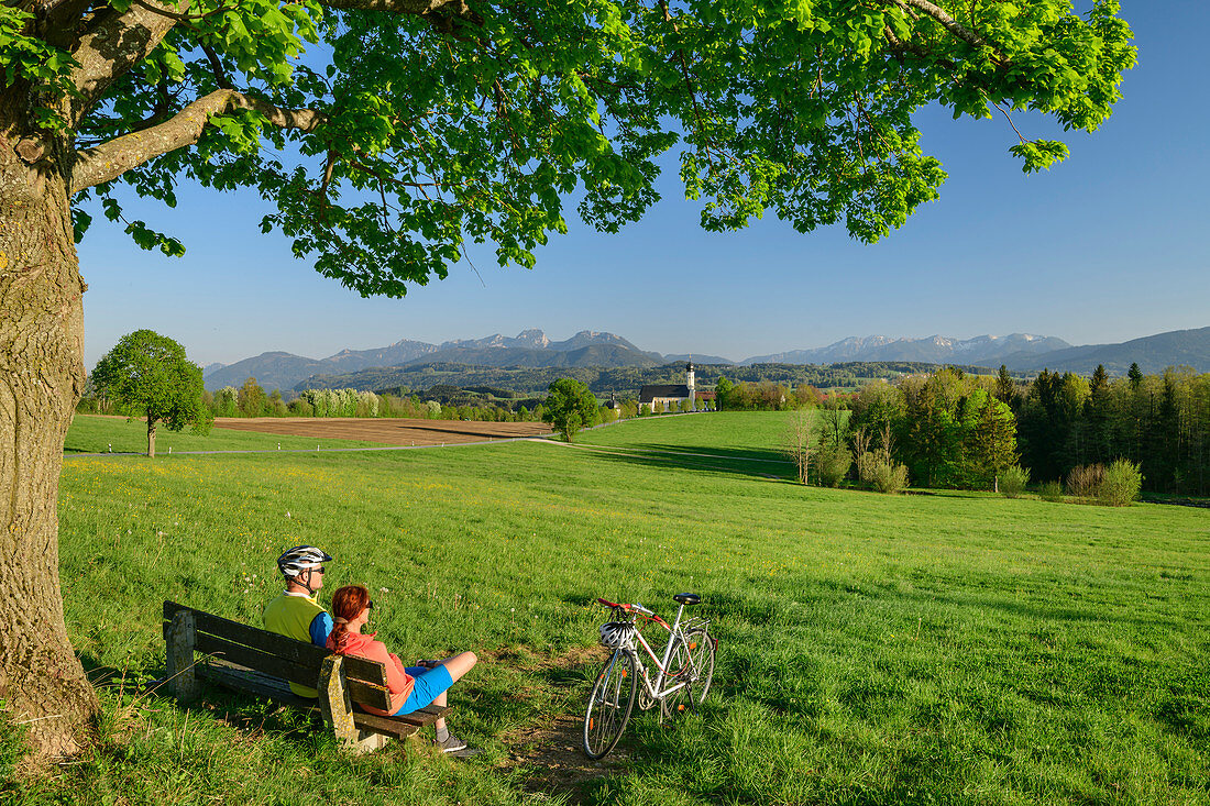 Frau und Mann beim Radfahren sitzen auf Bank und machen Pause, Kirche Wilparting und Mangfallgebirge im Hintergrund, Irschenberg, Oberbayern, Bayern, Deutschland