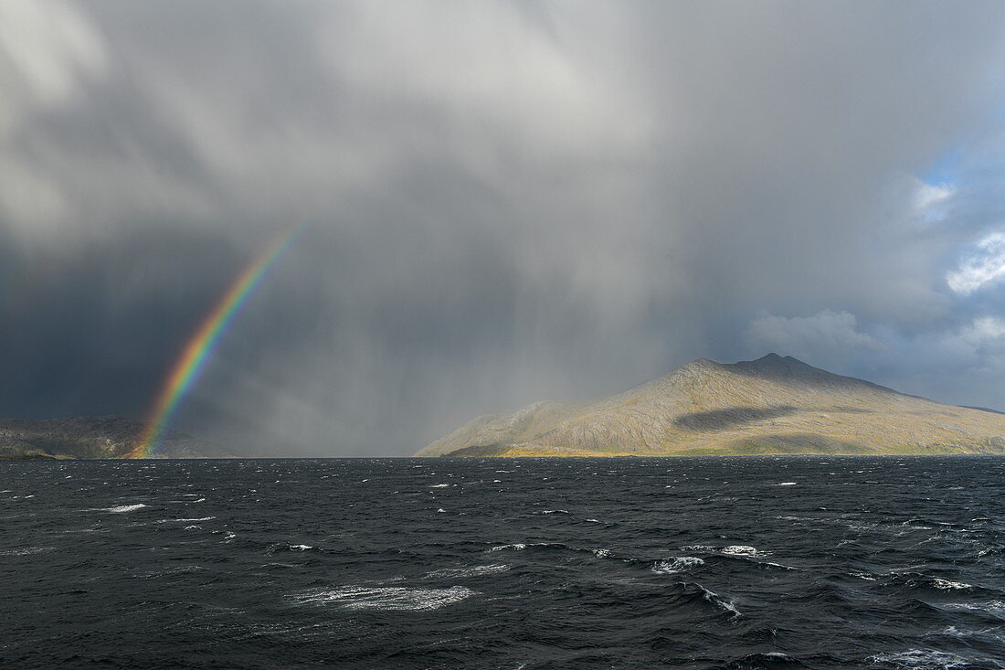 Ein Regenbogen bildet sich zwischen dicken Wolken, die über niedrige Berge treiben, chilenische Fjorde, Magallanes y de la Antartica Chilena, Patagonien, Chile, Südamerika