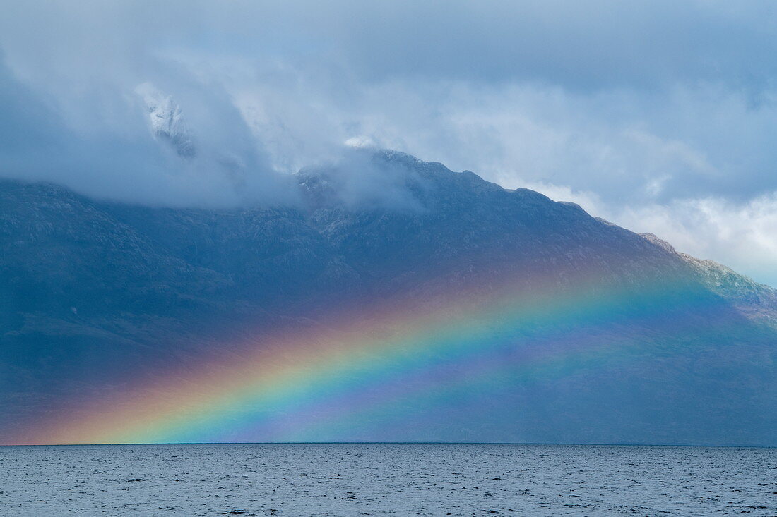 Ein heller Regenbogen bildet sich vor einem Berg, der von niedrigen Wolken umgeben wird, nahe Kirke Narrows, Magallanes y de la Antartica Chilena, Patagonien, Chile, Südamerika