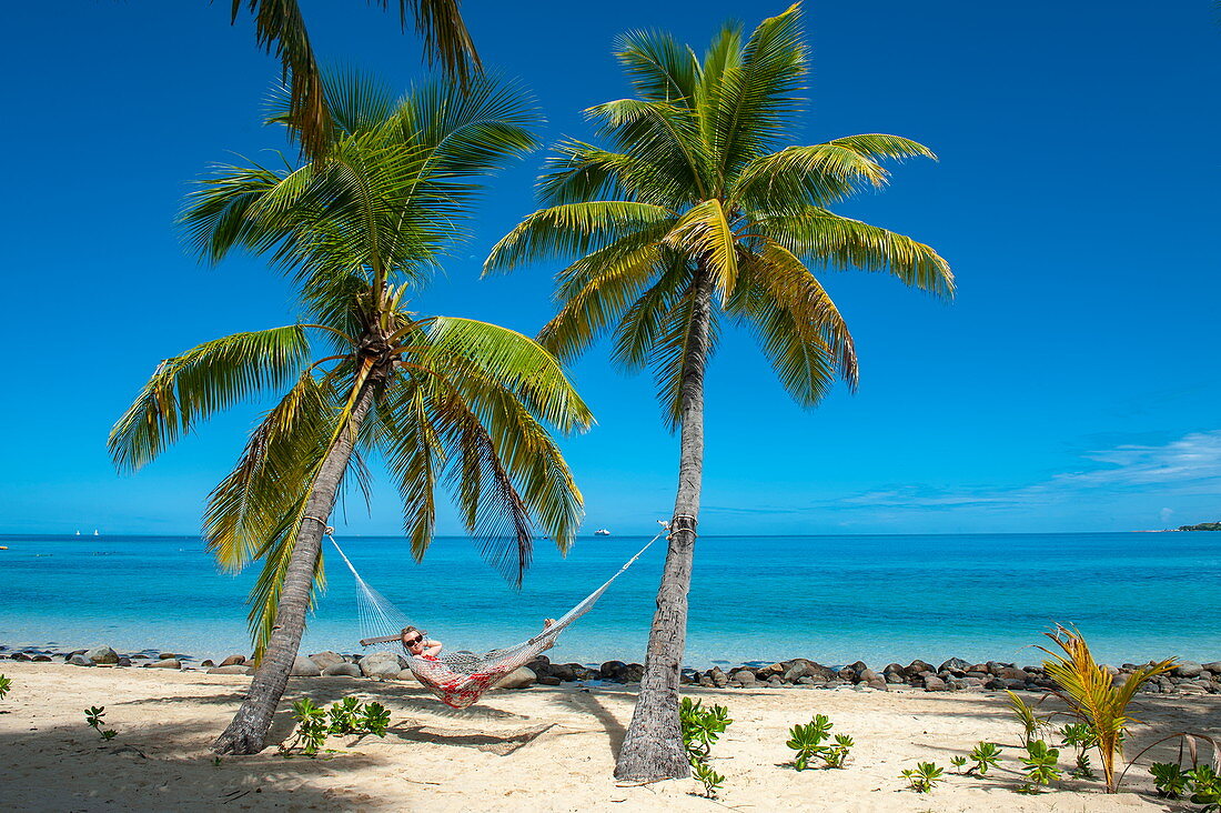 A woman enjoys the sun while lying in a hammock attached to two palm trunks near an idyllic beach, Mamanuca Islands, Fiji, South Pacific