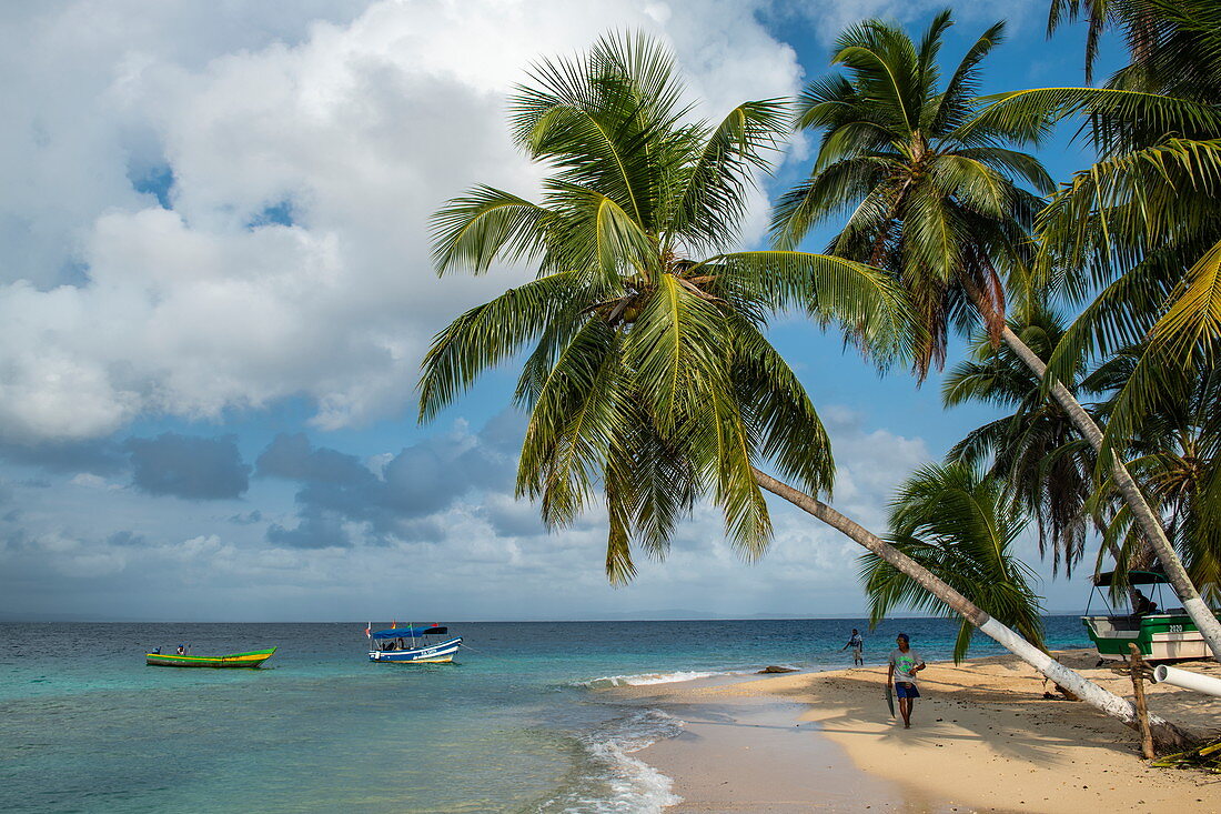 Palm trees line a narrow, sunny beach with boats and two people, San Blas Islands, Panama, Caribbean