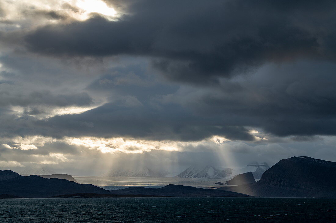 Landschaftsaufnahme der Sonne, die durch Wolkenöffnungen bricht, um auf eine karge, schneebedeckte Szene zu scheinen, Ny-Ålesund, Spitzbergen, Norwegen, Europa