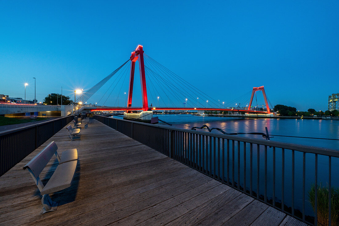 Illuminated, red Willemsbrücke during the blue hour, with plank path and seating on the river bank. Rotterdam, The Netherlands, June 2020