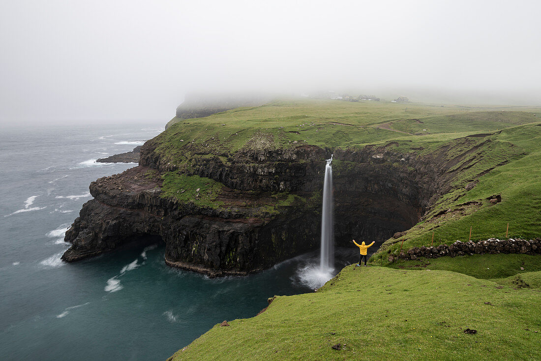 Hiker admiring the Mulafossur waterfall and the village of Gasadalur, Vagar island, Faroe Islands, Denmark, Europe