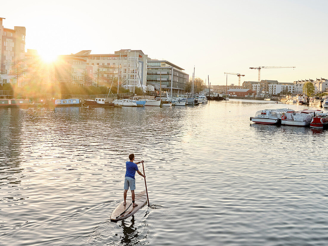 Man standing on paddleboard on river at dawn, shot from behind