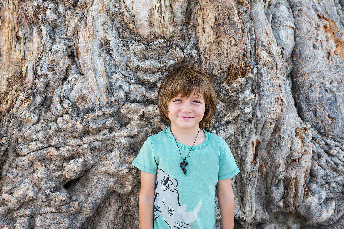 A five year old boy posing against trunk of a large Baobab tree