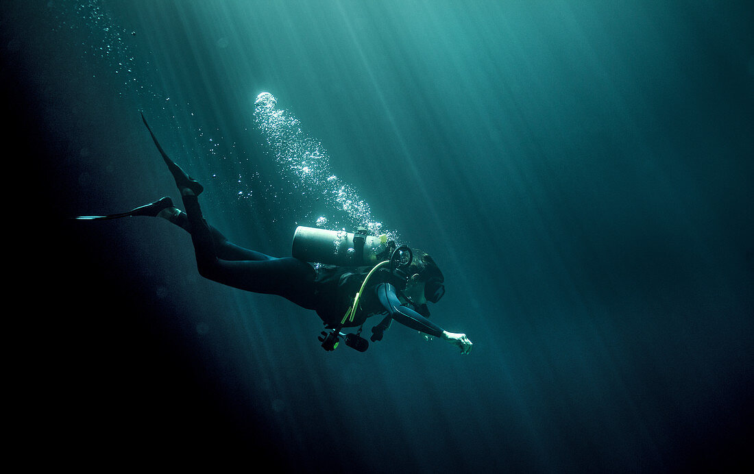 Underwater view of diver wearing wetsuit, diving goggles and oxygen cylinder, air bubbles rising.