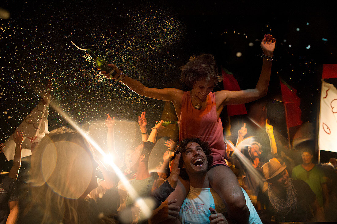 Revellers at an open air concert, smiling man carrying woman on his shoulders, arms outstretched, holding beer bottle.