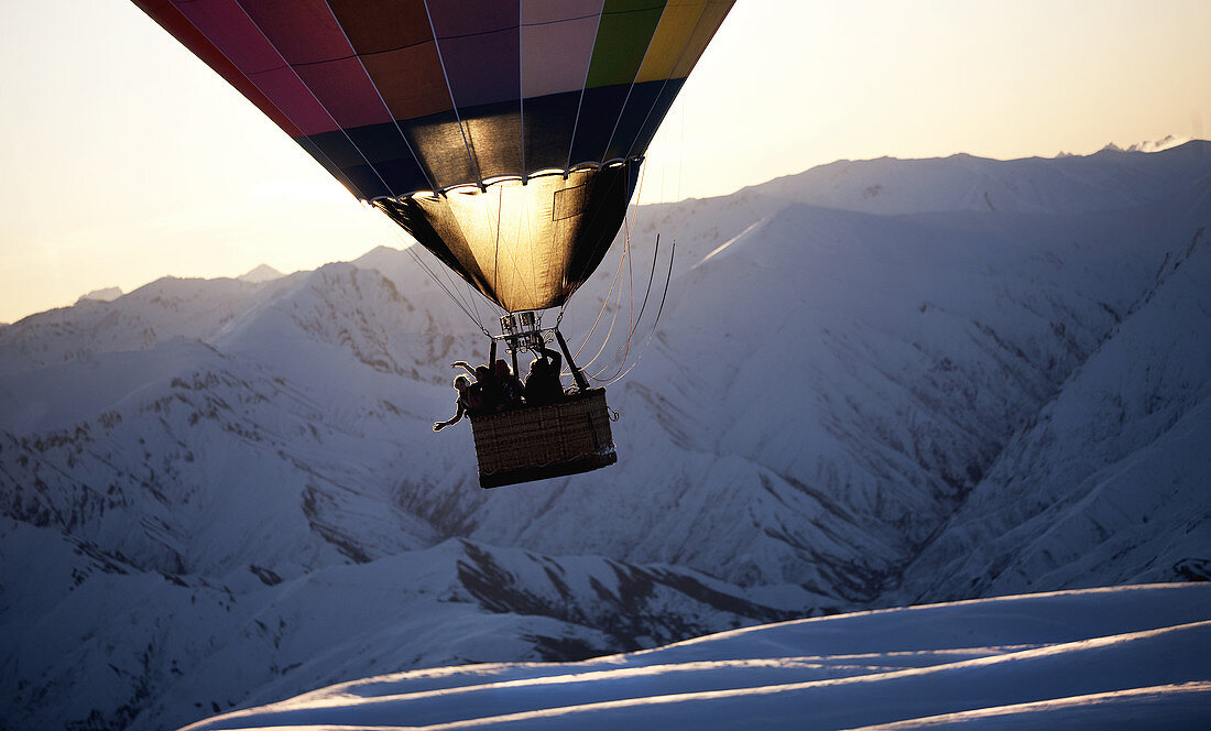 People in a hot air balloon mid air over a snow covered mountain.