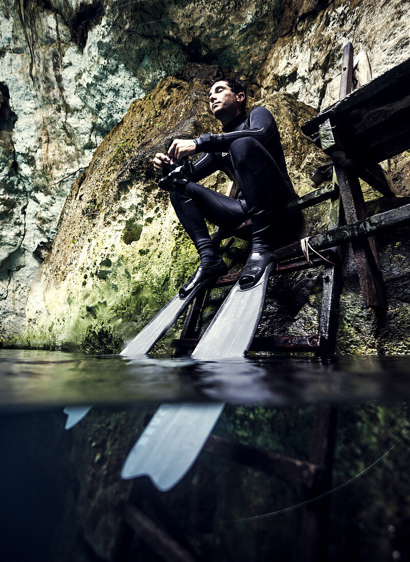 Man wearing wet suit and flippers sitting on a platform, rocks in background.