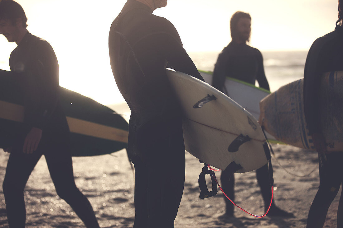 Four men wearing wetsuits standing on a sandy beach, carrying surfboards.