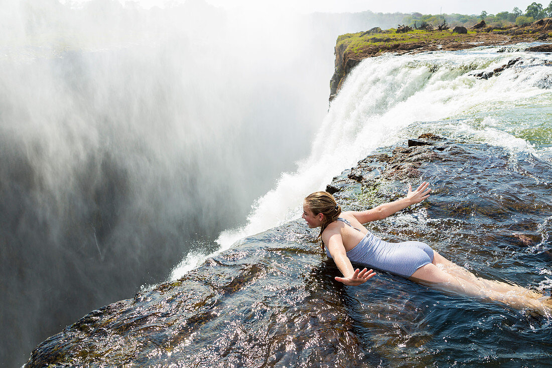 Young girl in the water at the Devils Pool lying on her front, arms spread out, at the edge of the cliff of Victoria Falls.