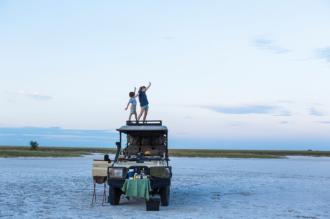 Bruder und Schwester stehen auf Safari-Jeep in der Dämmerung in einer Salzpfannen-Landschaft