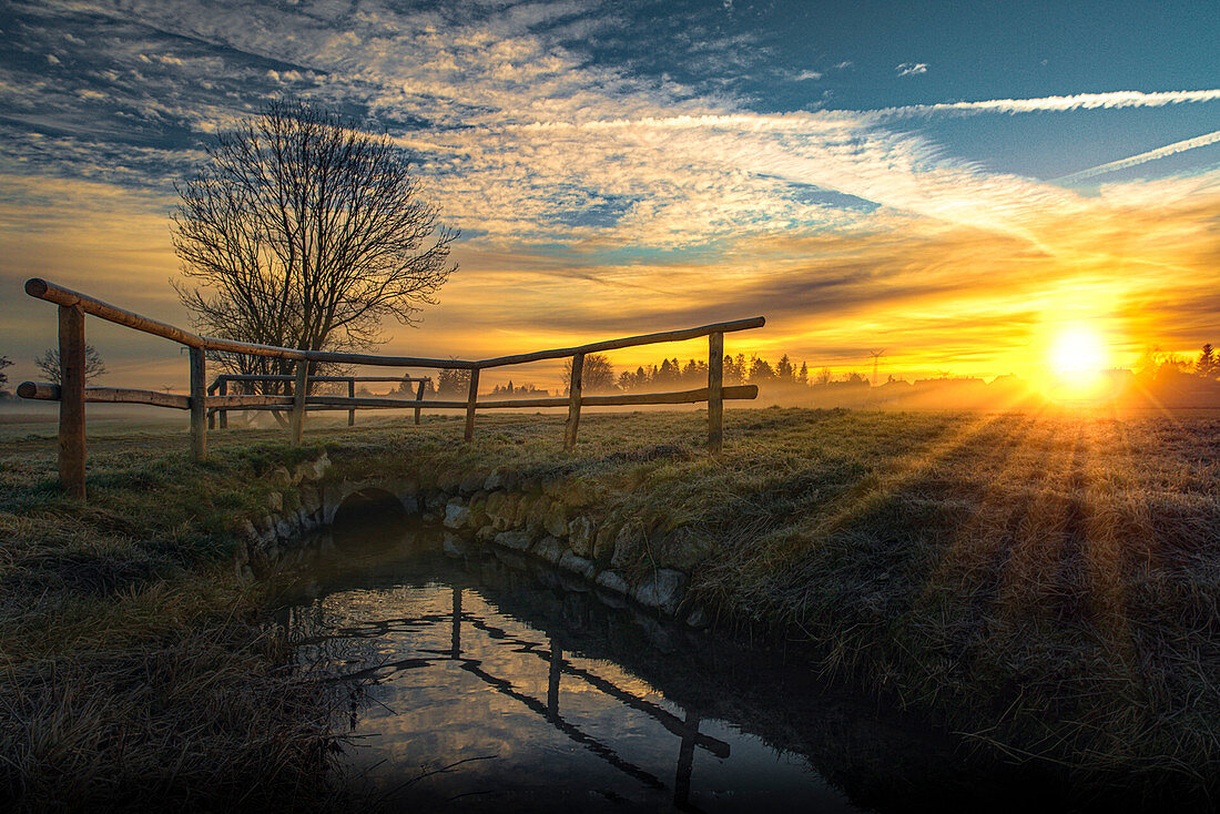 Small brook with bridge and rising sun on a field in the west of Munich. Munich, Upper Bavaria, Bavaria, Germany, Europe
