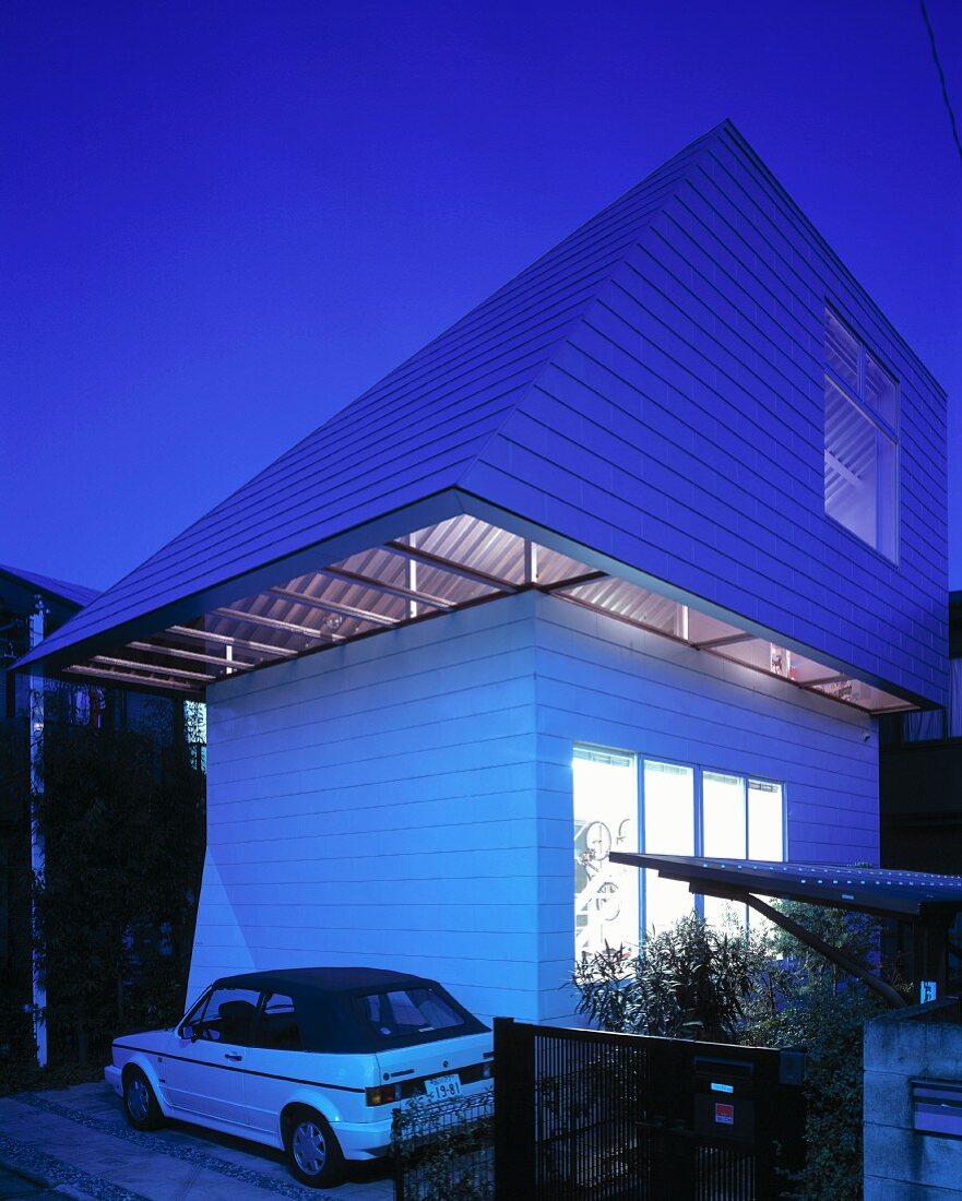 A parked car in front of a newly built house with a white facade and a green slate roof in the evening
