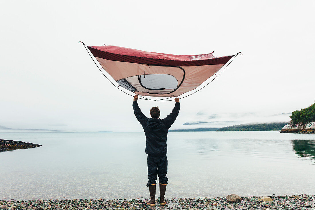 Man holding camping tent over head, standing on rocky beach,an inlet on the Alaska coastline.