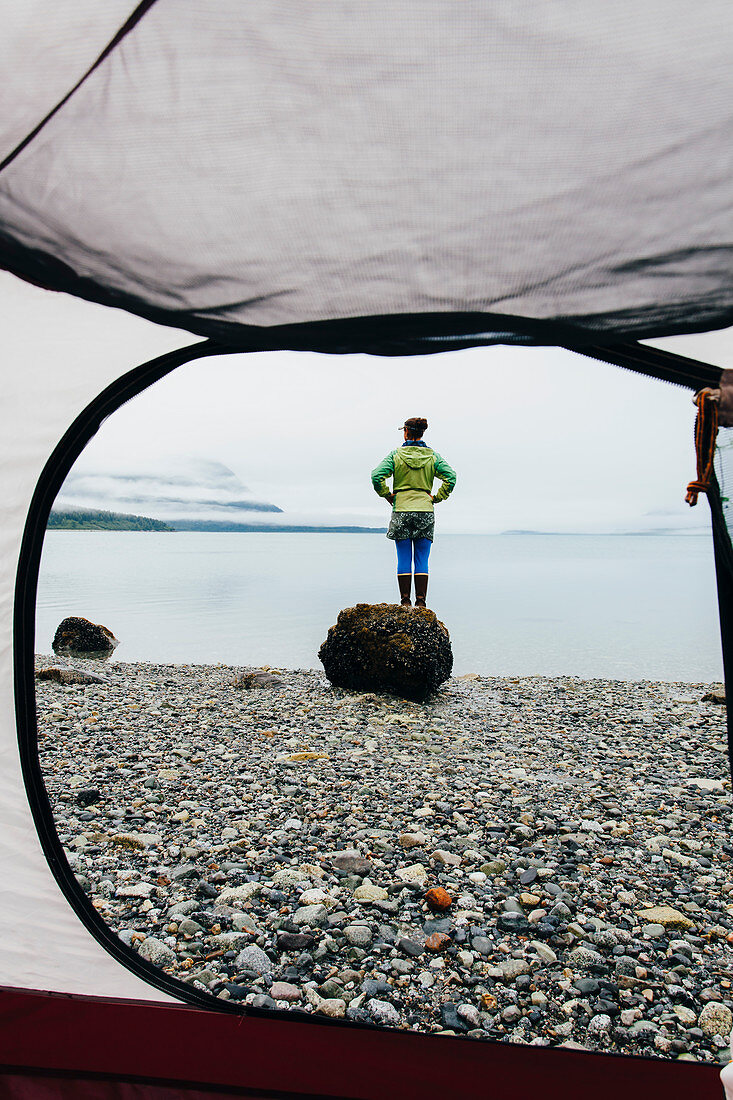 View through camping tent doorway of woman standing on beach,an inlet on the Alaska coastline.
