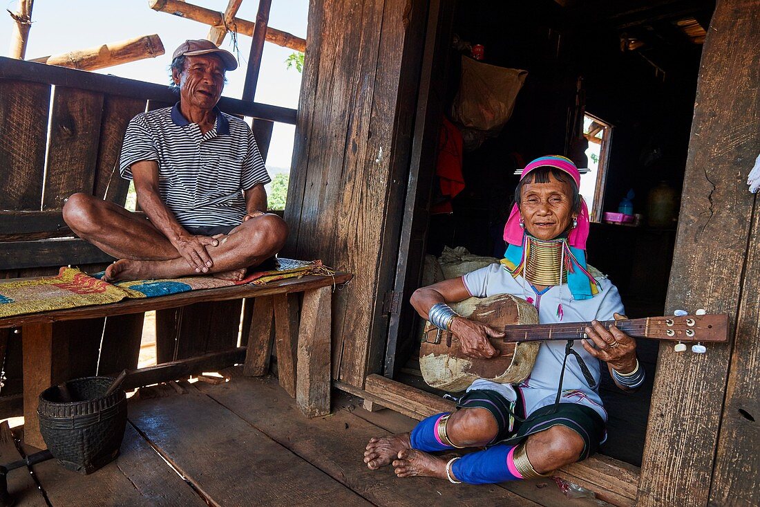Kayan Lahwi Frau mit Messinghalsringen und traditioneller Kleidung, die Gitarre auf ihrem Hausdeck spielt. Ihr Mann hört zu. Pan Pet Region, Bundesstaat Kayah, Myanmar.