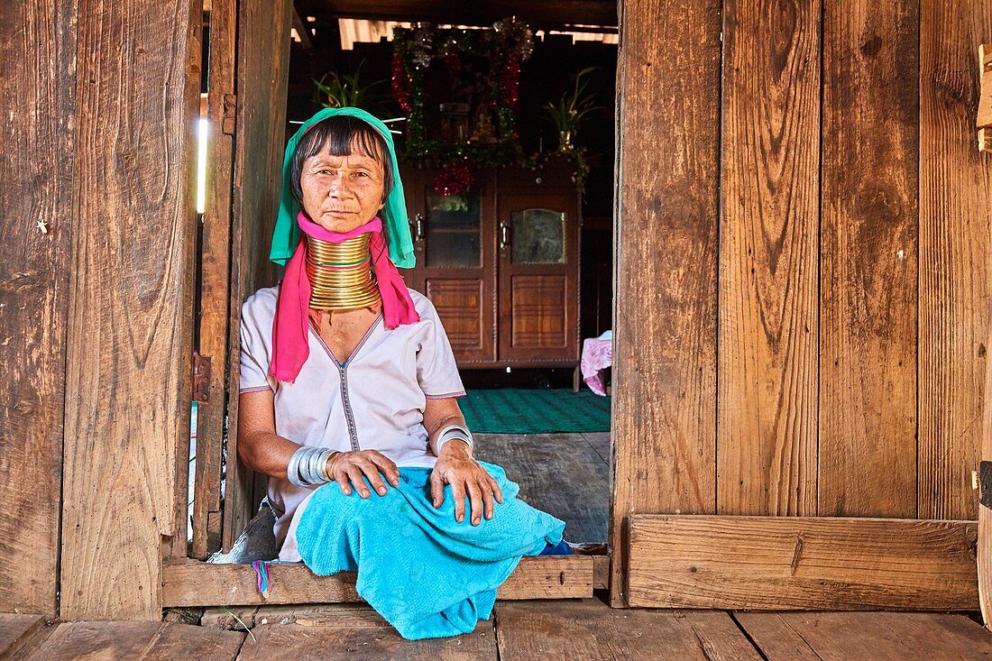 Kayan Lahwi Frau mit Messinghalsringen und traditioneller Kleidung sitzt neben der Haustür ihres Hauses. Im Hintergrund steht ein buddhistischer Altar.  Pan Pet Region, Bundesstaat Kayah, Myanmar.
