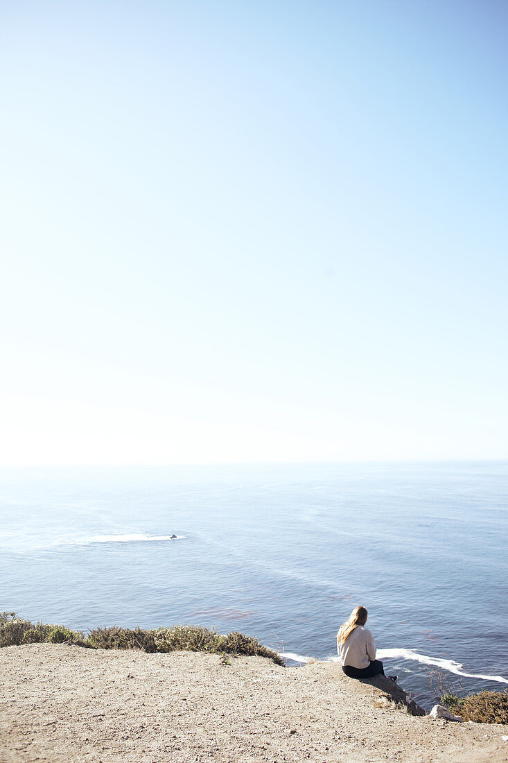 Young woman sits on a cliff and looks out over the Pacific Ocean on the edge of Highway 1, Big Sur State Park, California, USA.