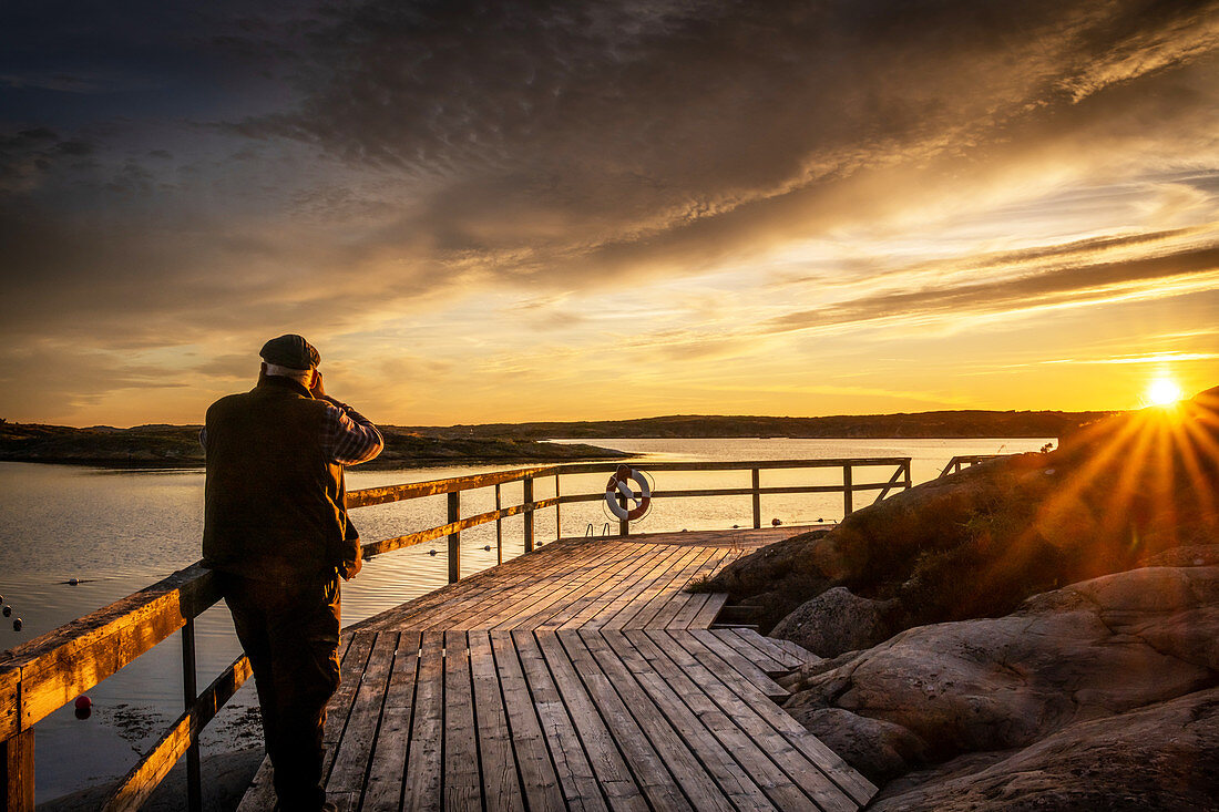 Ein Mann beobachtet den Sonnenuntergang auf einem Steg in den Schären, Ellös, Orust, Bohuslän, Schweden