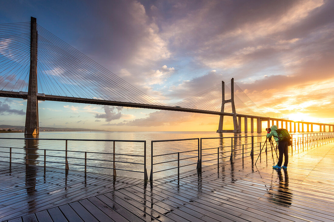 A photographer taking pictures on a pier during a colorful sunrise in front of the Vasco da Gama bridge. Lisbon, Portugal, Europe.