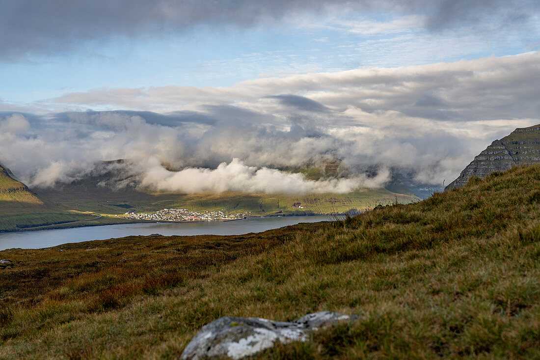 Blick vom Klakkur auf das Dorf Leirvík mit sonnendurchschienen Wolken und der typischen Graslandschaft, Färöer Inseln