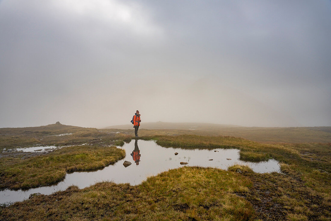 Hiker stands on a mirror-smooth water surface in the high moor of the Klakkur, Klaksvík, Faroe Islands.