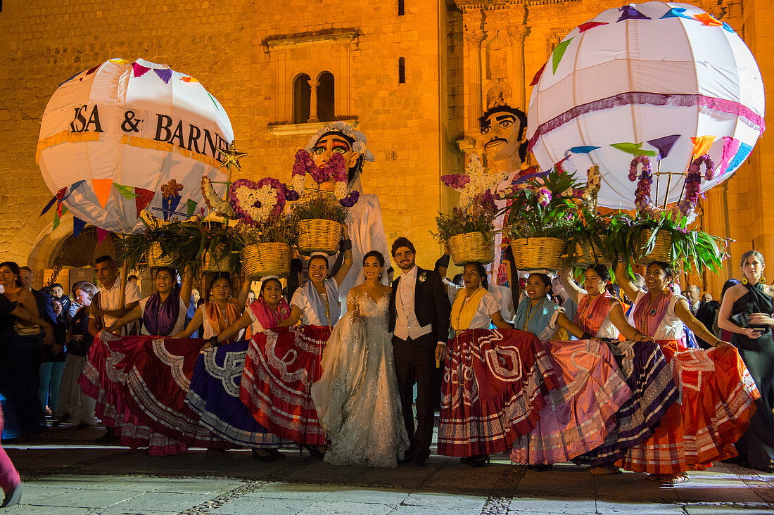 Women dressed in a regional costume and giant puppets dressed as a bride and groom during a Calenda, a procession on the Plaza Santo Domingo, celebrating a wedding in the city of Oaxaca de Juarez, Oaxaca, Mexico.