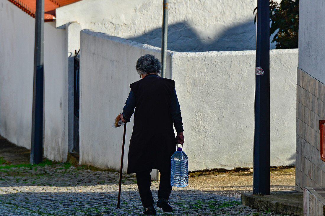 Eine alte Frau kommt vom Einkauf und geht durch die Dorfstraße, Odeceixe, Algarve, Portugal