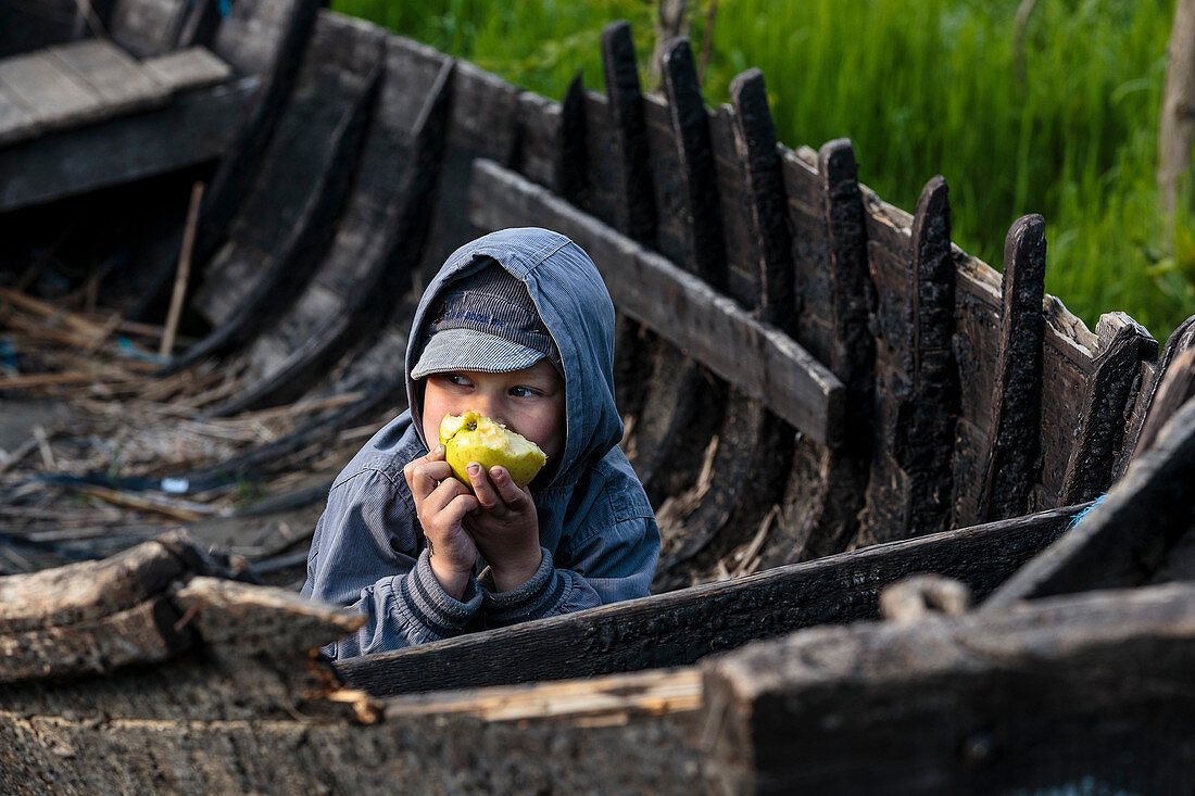 Ein Junge isst einen Apfel In einem verfallenen Holzboot, Donaudelta im April, Mila 23, Tulcea, Rumänien.