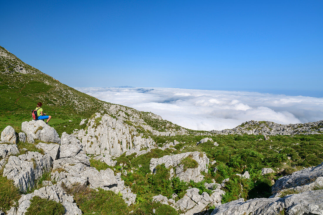 Frau beim Wandern sitzt auf Felsen und blickt auf Nebelmeer, vom Picu Tiedu, Nationalpark Picos de Europa, Kantabrisches Gebirge, Asturien, Spanien