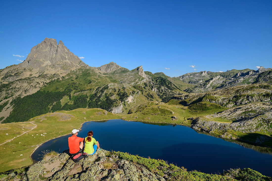 Mann und Frau beim Wandern sitzen auf Felssporn und blicken auf Lac Roumassot und Pic du Midi d´Ossau, Lac Roumassot, Nationalpark Pyrenäen, Pyrénées-Atlantiques, Pyrenäen, Frankreich