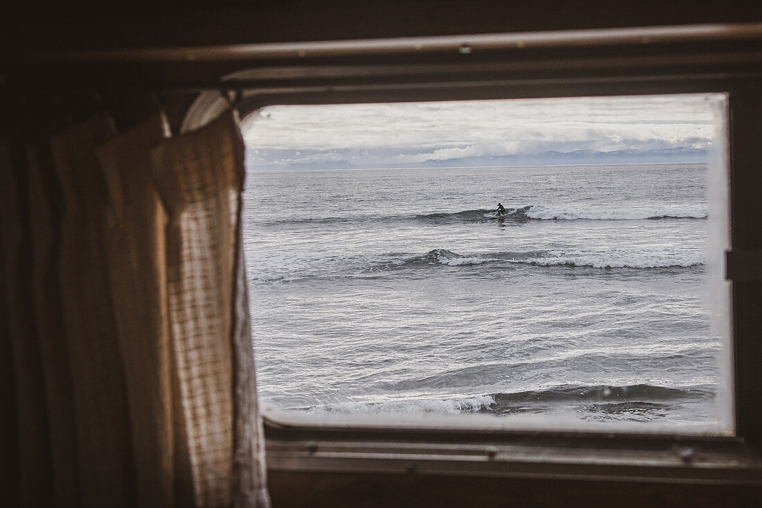 View of ocean through camper van window, surfer in the distance.