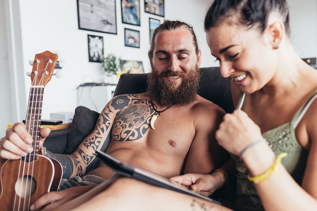 Bearded tattooed man with long brunette hair and woman with long brown hair sitting on a sofa, looking at digital tablet.